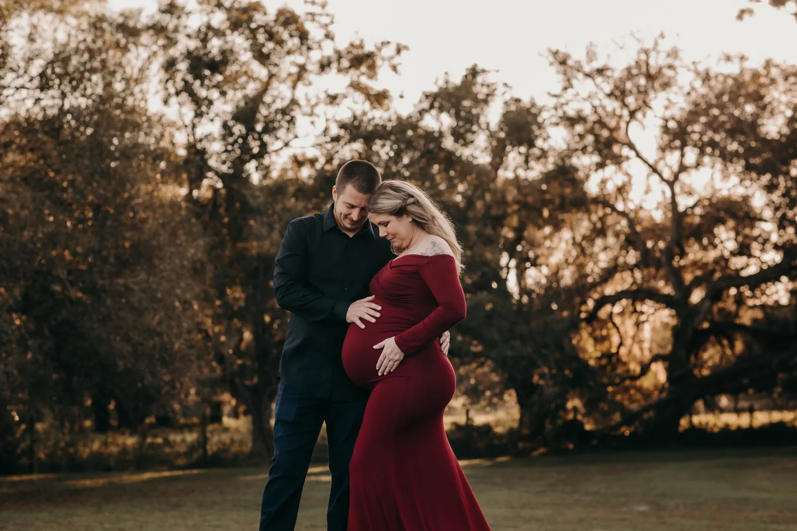 Couple with their heads together and hands on her bump, in front of the oaks