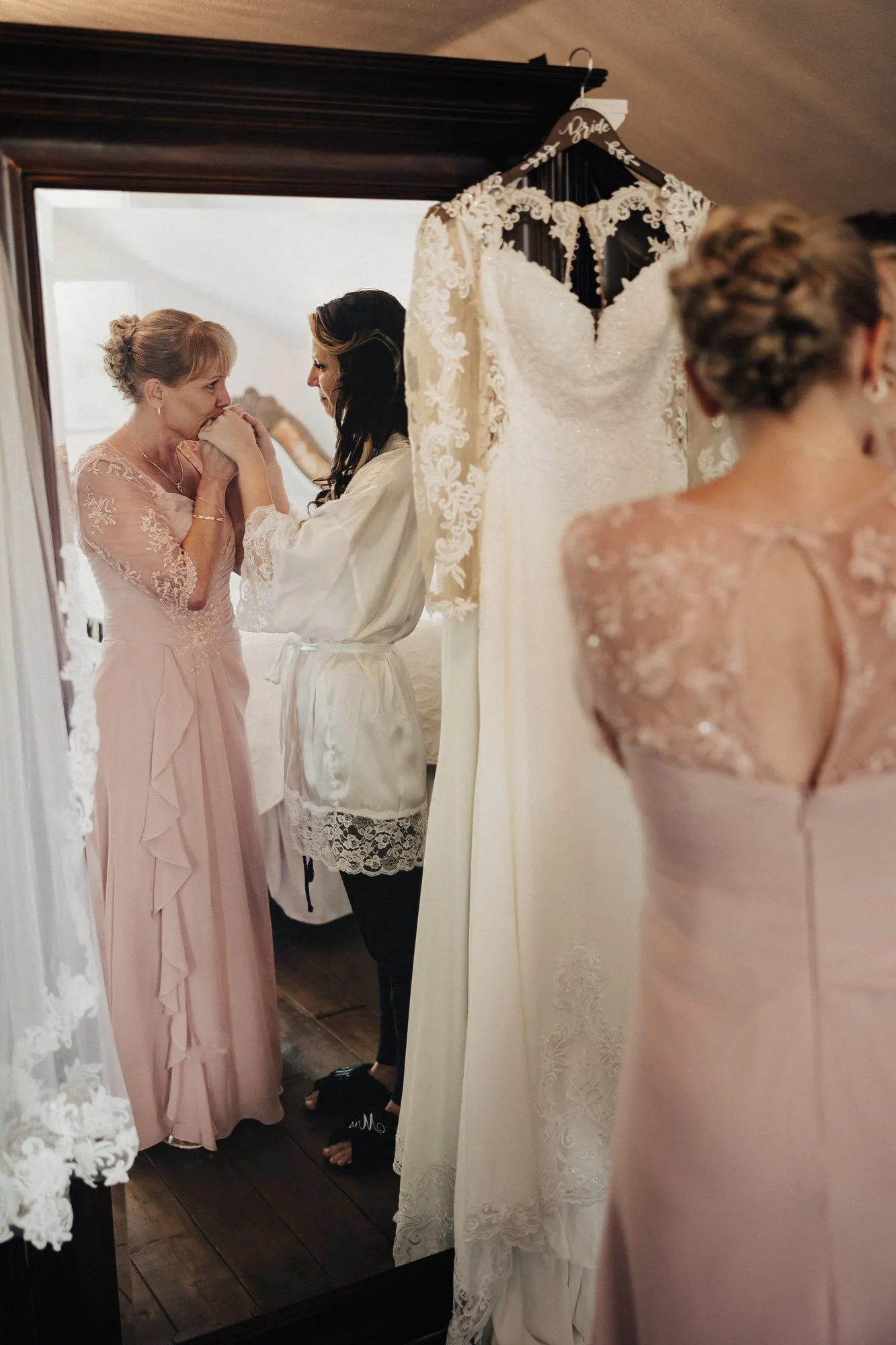 Woman in a blush dress kissing the bride's hands beside her hanging gown
