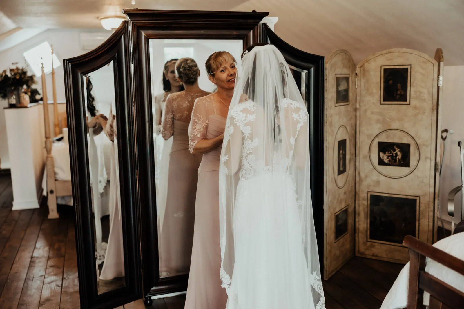 Bride in her veil, reflected in a mirror, as a woman fastens her dress