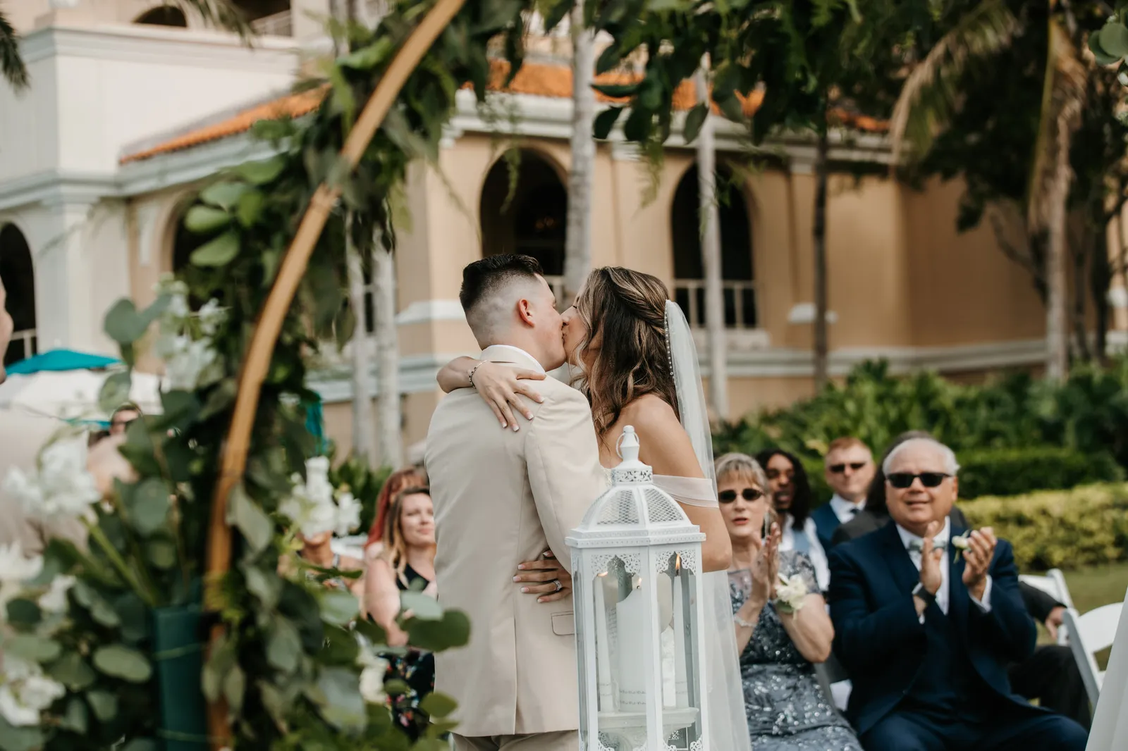 Bride and groom's first kiss under a floral arch as guests applaud