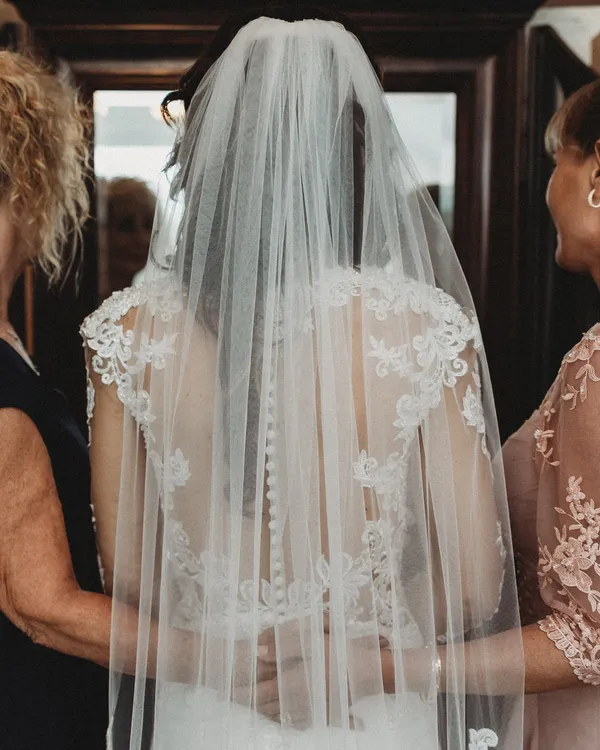 Bride in her veil, seen from behind, with two women helping her get ready