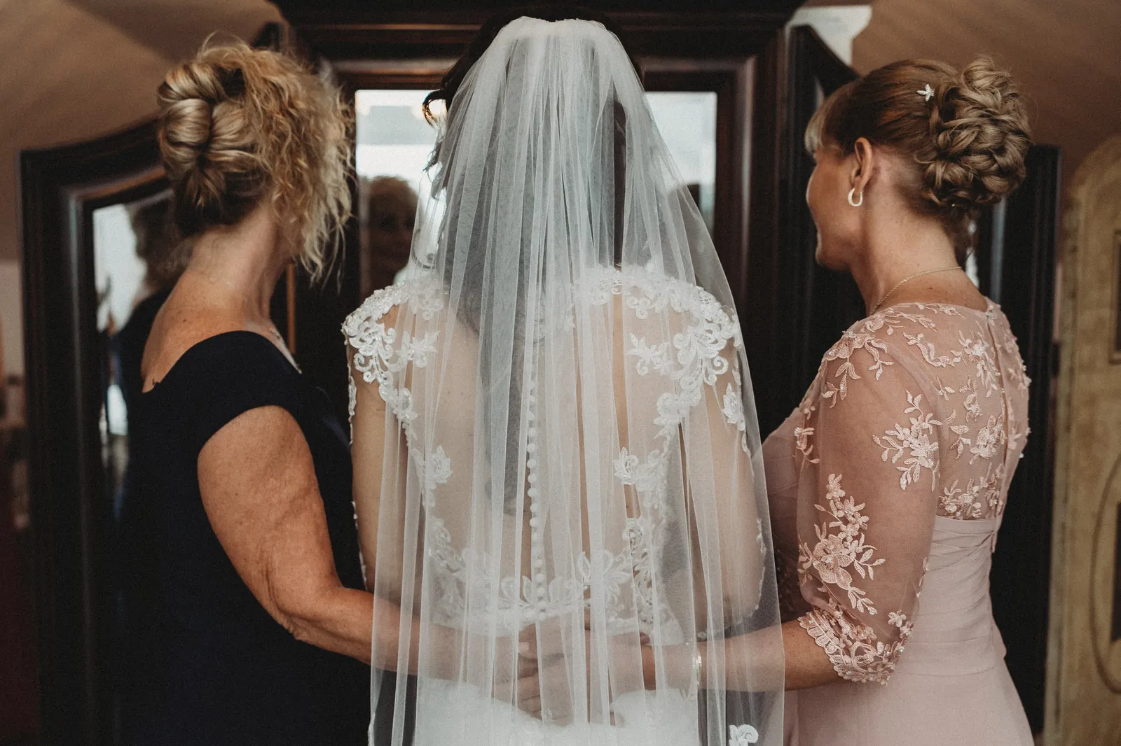 Bride in her veil, seen from behind, with two women helping her get ready