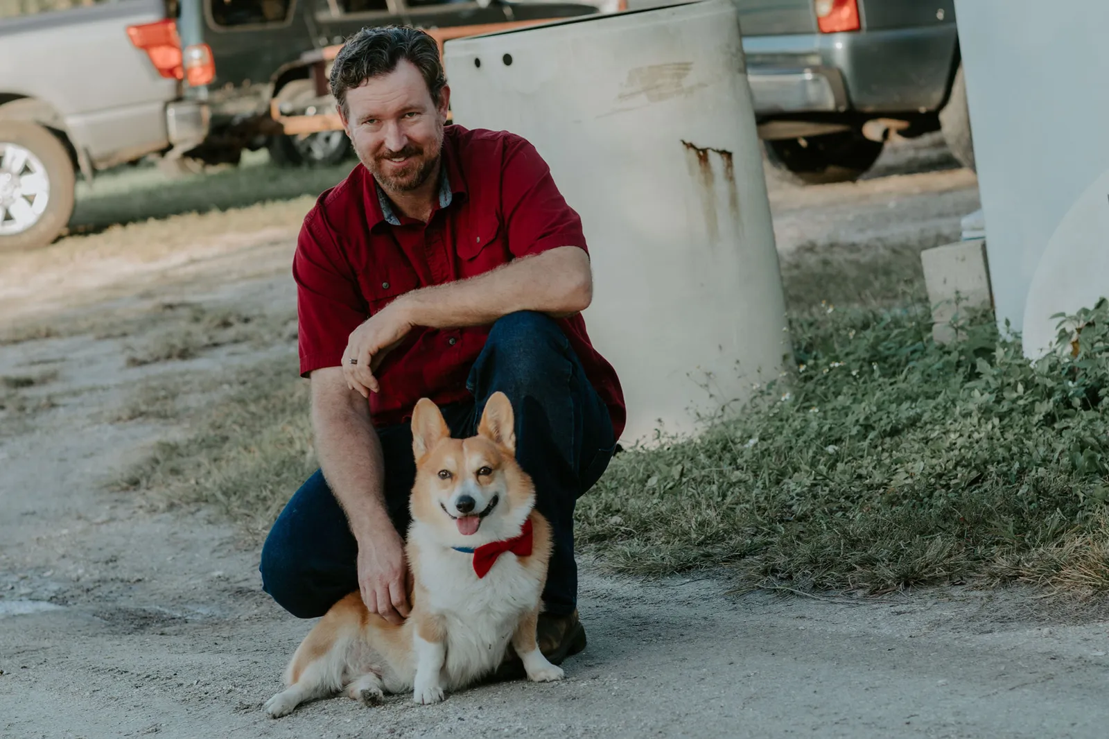 Man in a red shirt crouching with a corgi in a red bow tie