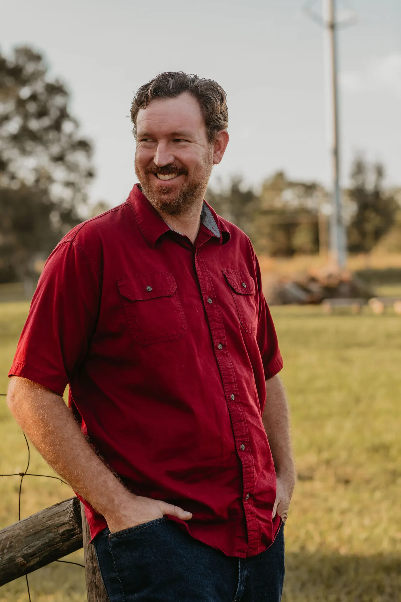 Man in a red shirt smiling in a field, hands in his pockets