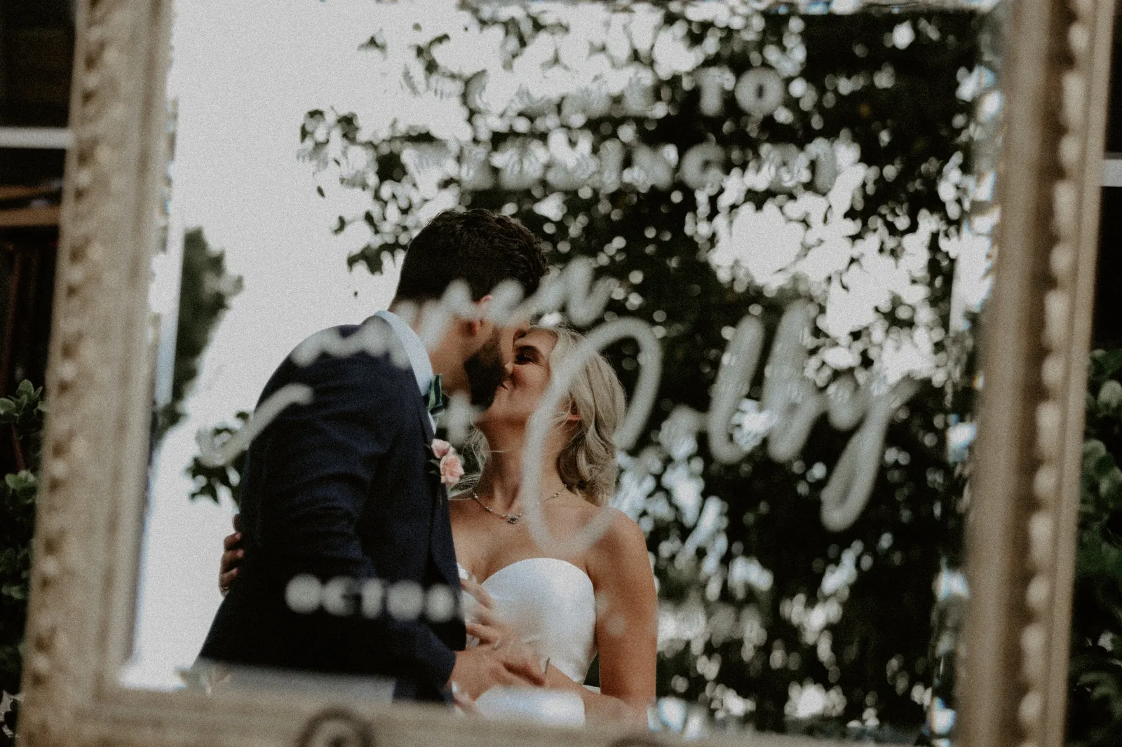 Bride and groom kissing, reflected in a gold-framed welcome mirror