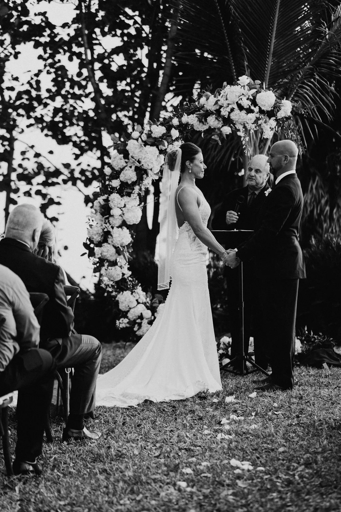 Bride and groom holding hands before the officiant under a floral arch, in black and white