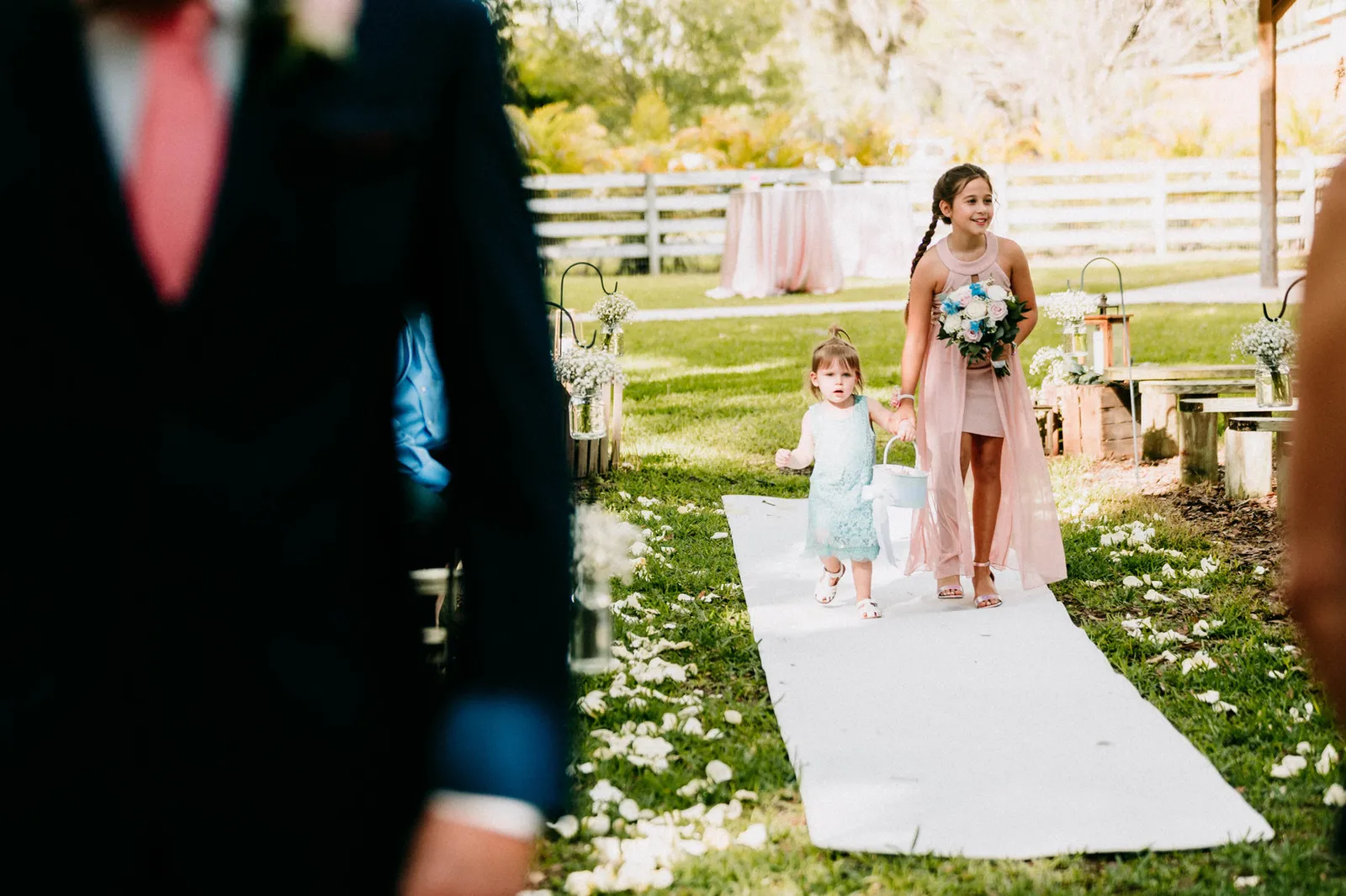 Junior bridesmaid and toddler flower girl on the white aisle runner, seen past the groom