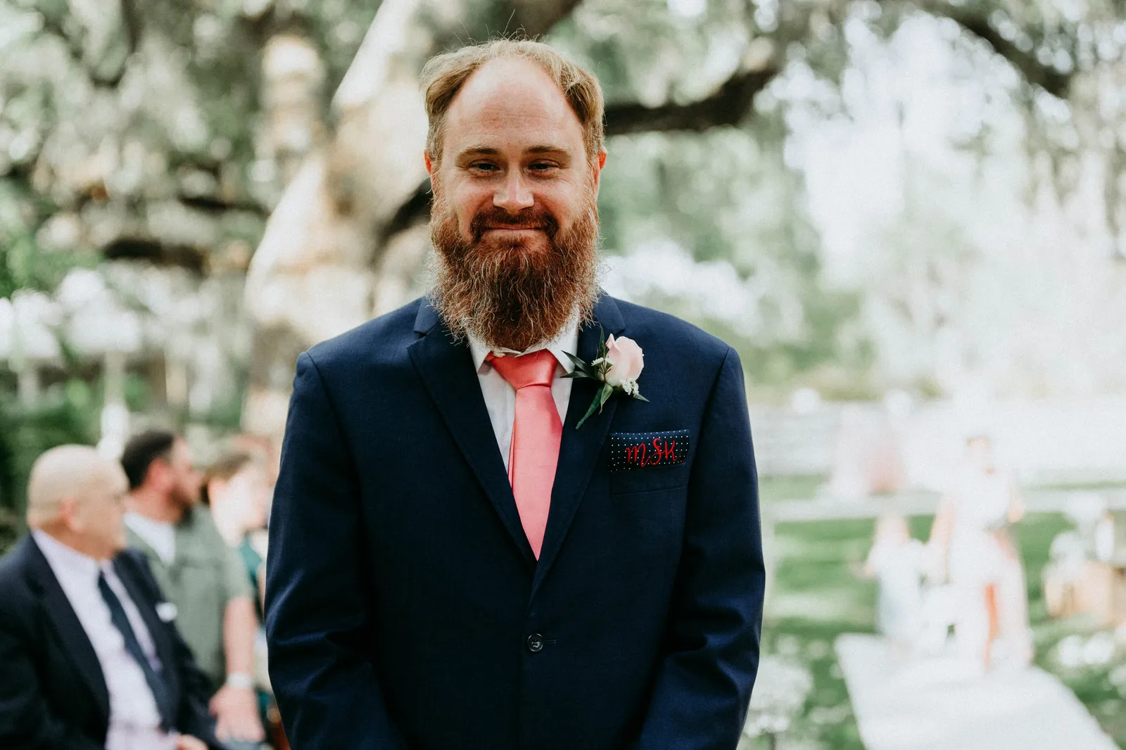 Bearded groom in a navy suit and coral tie smiling as he waits under the oaks