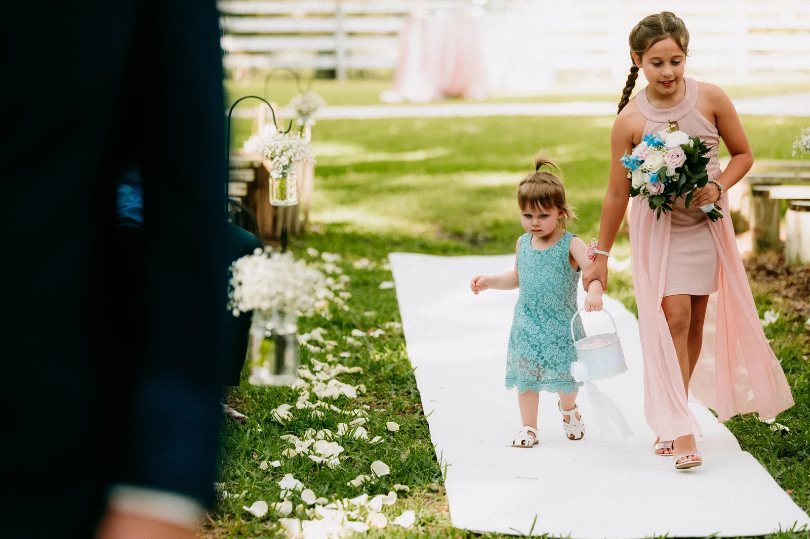Toddler flower girl in a teal dress walking the aisle beside a girl in pink