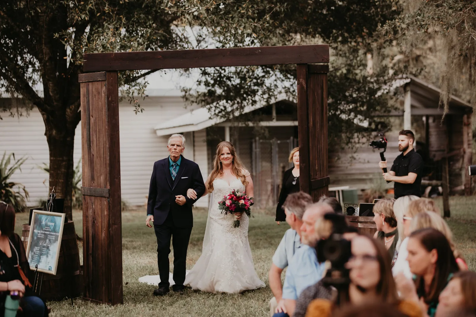 Bride walking toward a wooden arch on an older man's arm as guests turn to watch