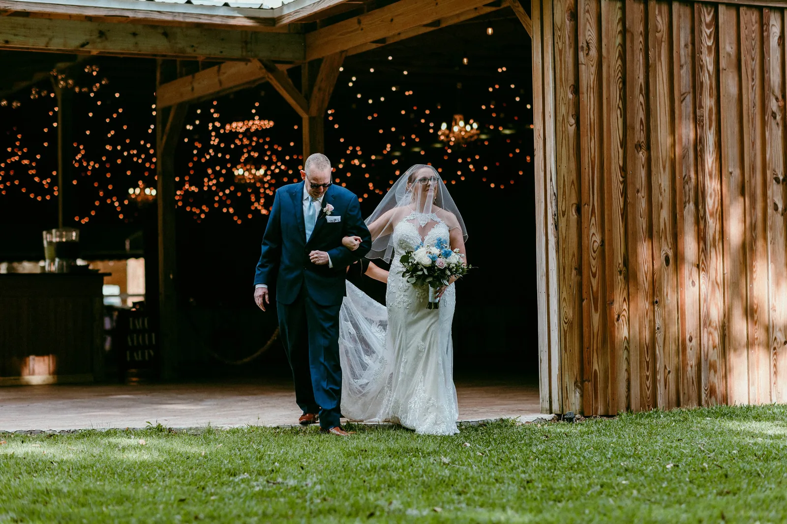 Bride walking out of the string-lit barn on the arm of a man in a suit