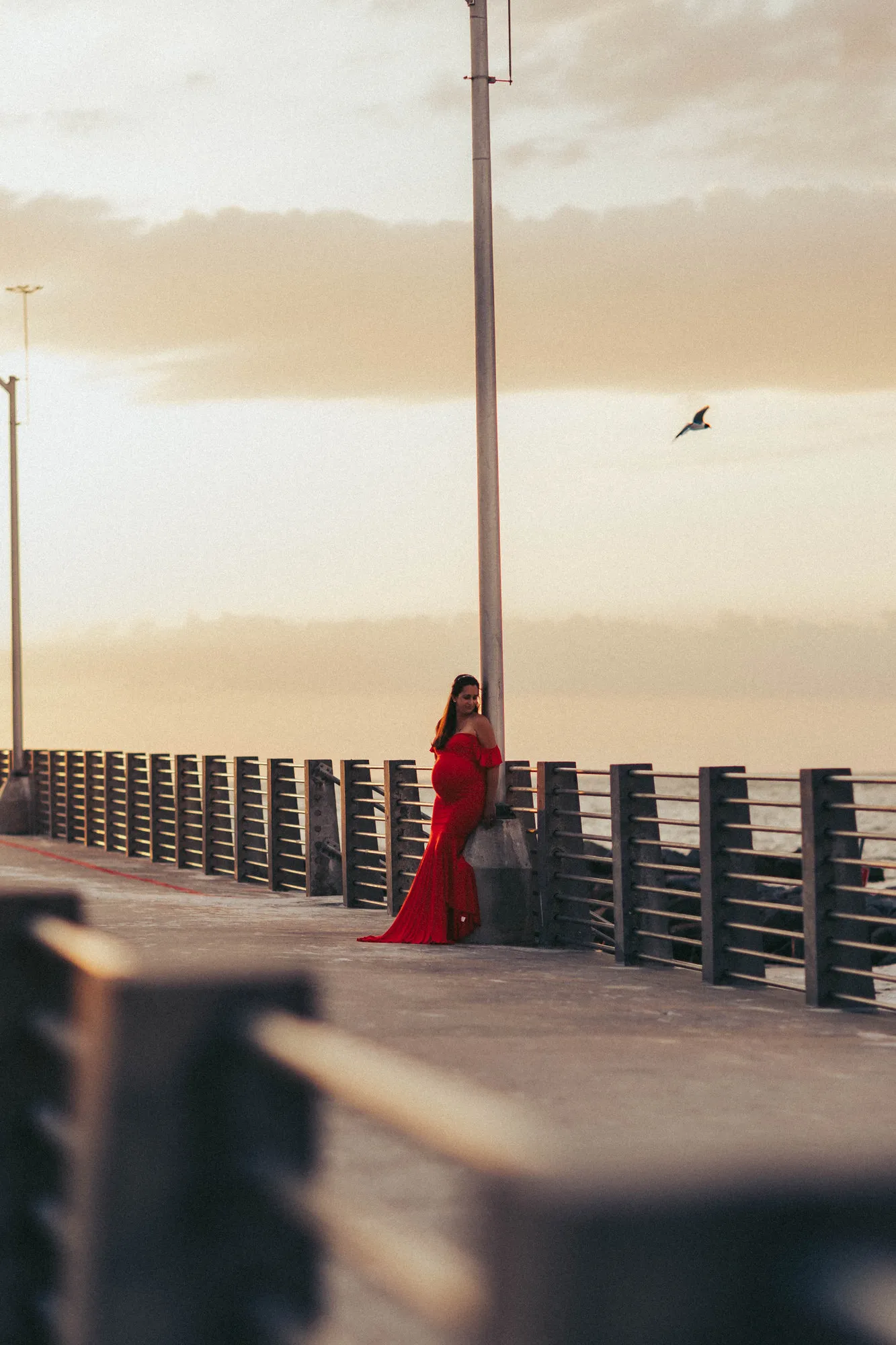 Expectant mother in a red gown leaning on the pier railing at sunset