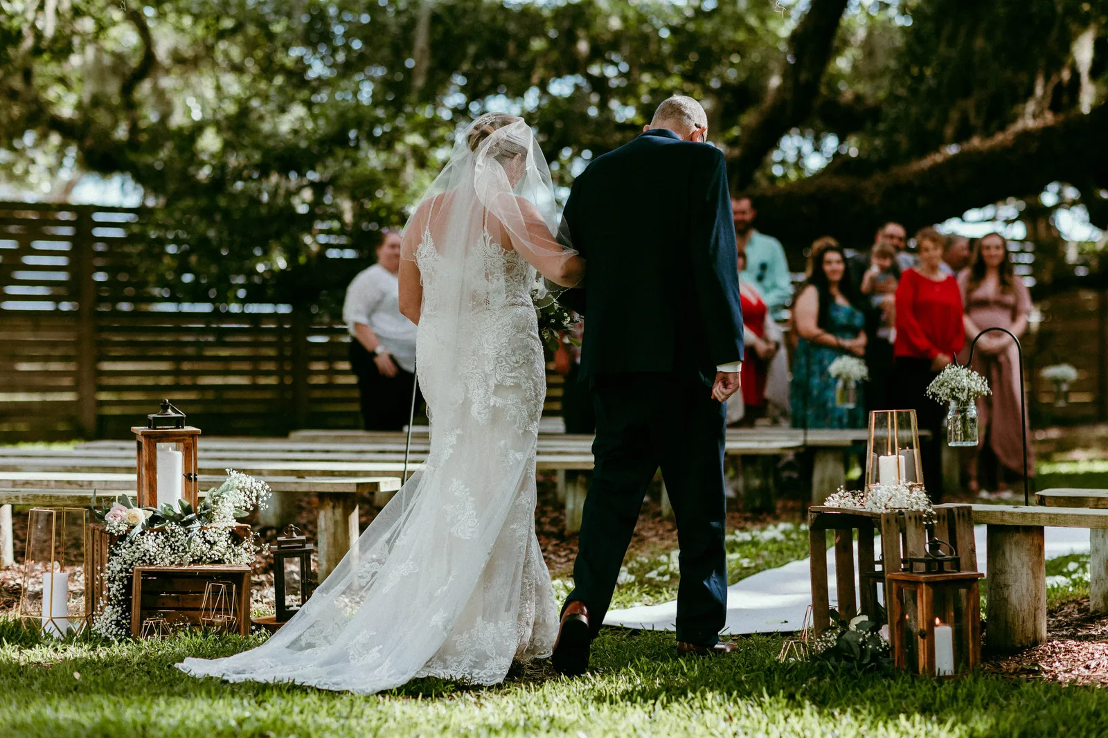 Bride being walked down the aisle beneath the oaks, seen from behind