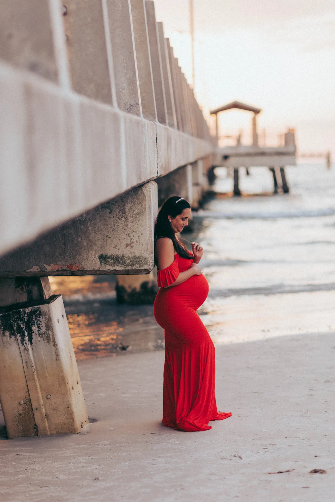 Expectant mother in a red gown beneath the pier at sunset