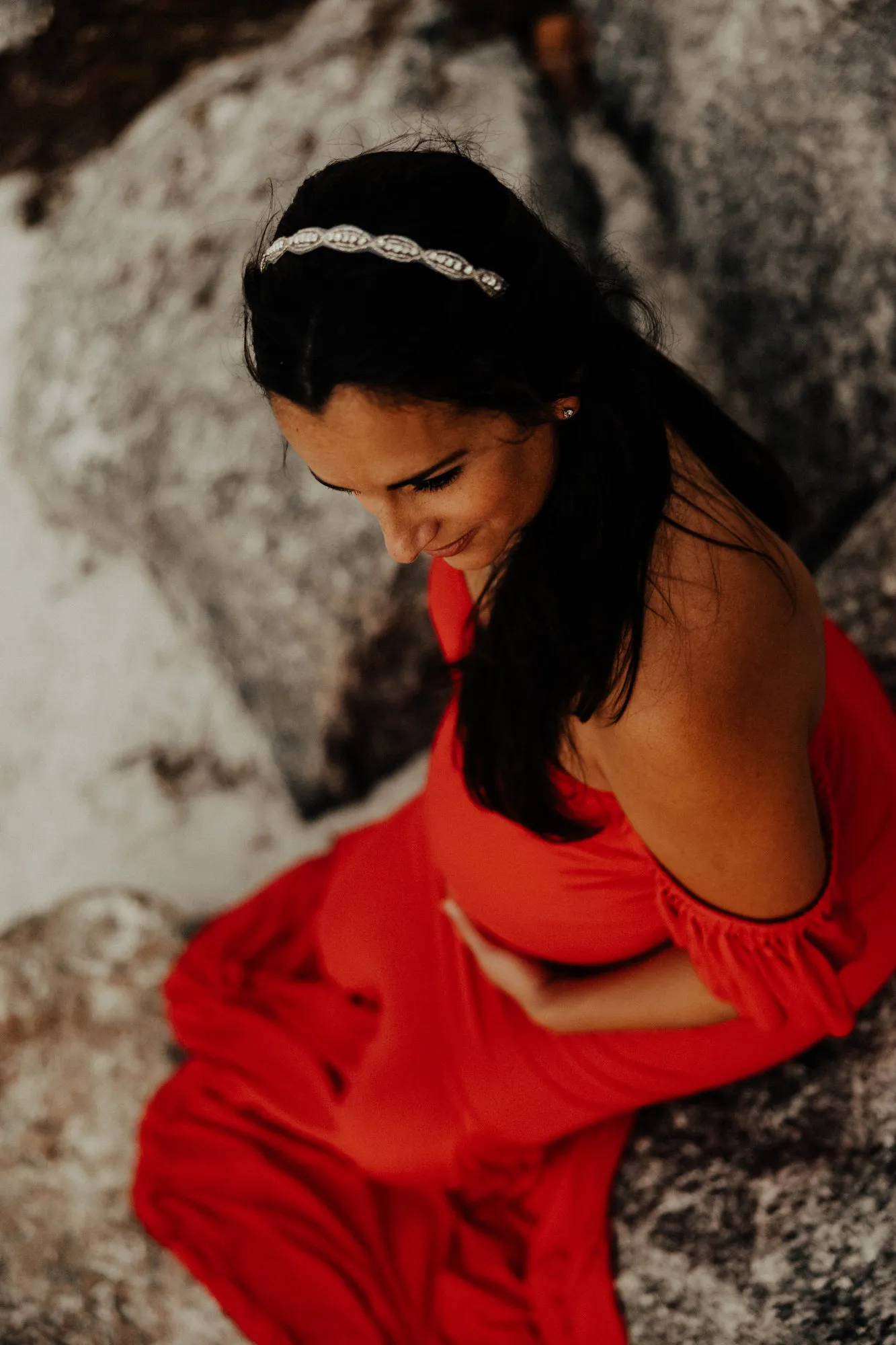 Expectant mother in a red gown and beaded headband sitting on the rocks, looking down