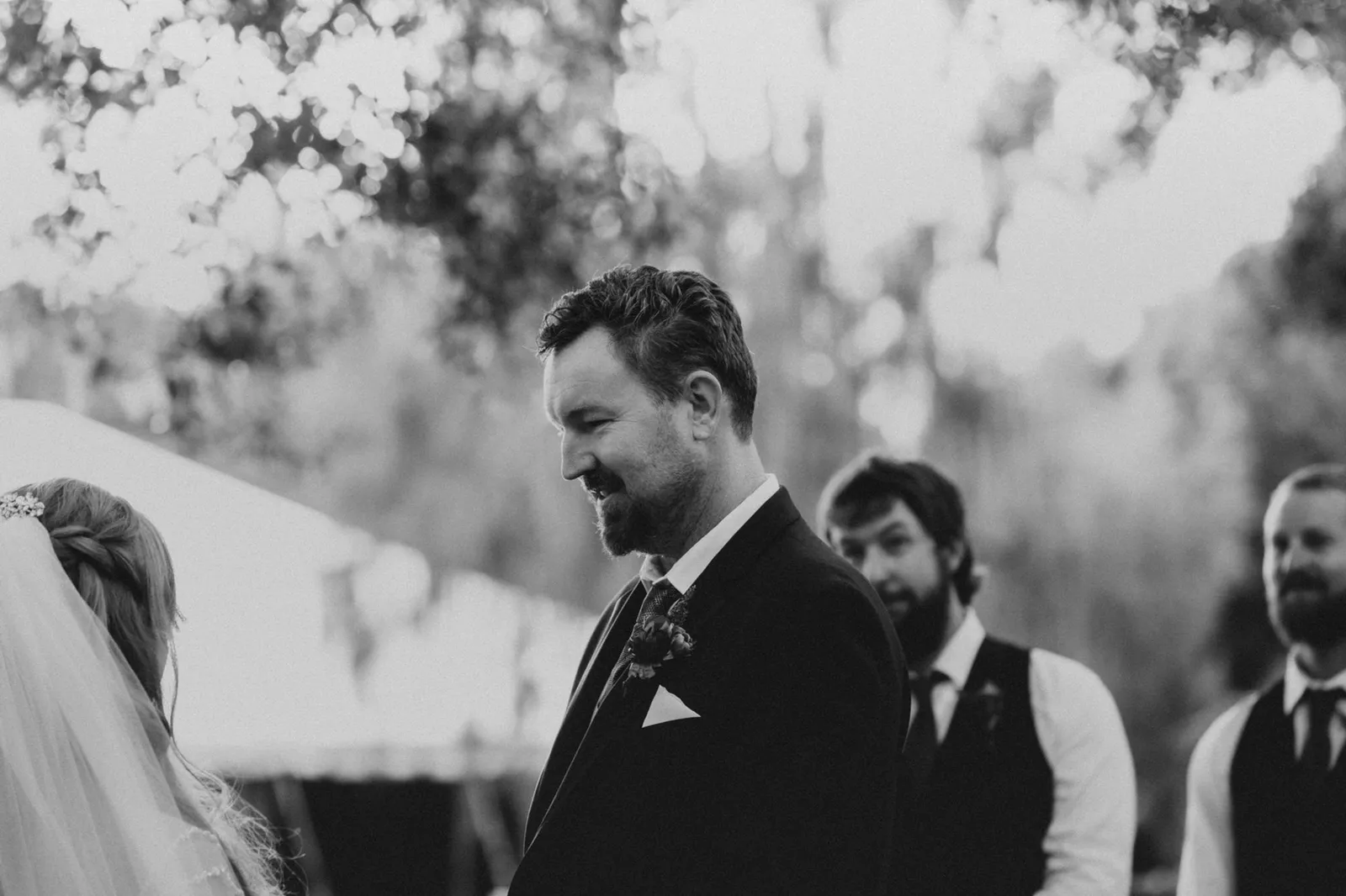 Groom smiling during the ceremony with his groomsmen behind him, in black and white