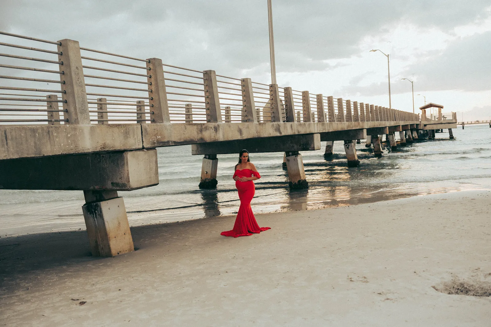 Expectant mother in red standing on the sand beside the Fort De Soto pier