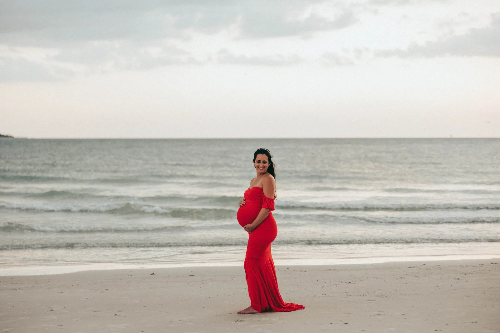 Expectant mother in a red gown at the water's edge under a cloudy sky