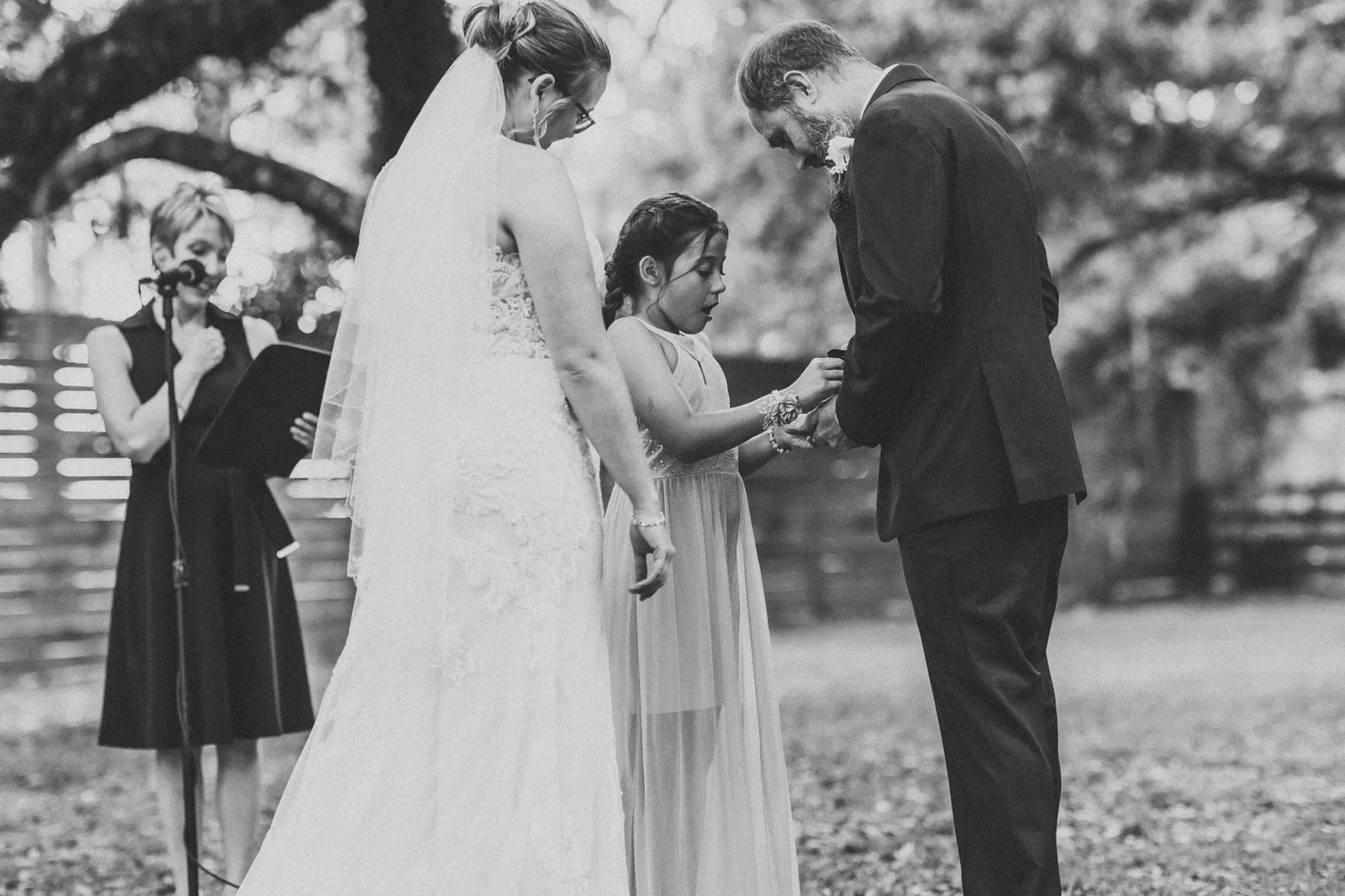 Groom fastening something to the girl's wrist as the bride and officiant look on, in black and white