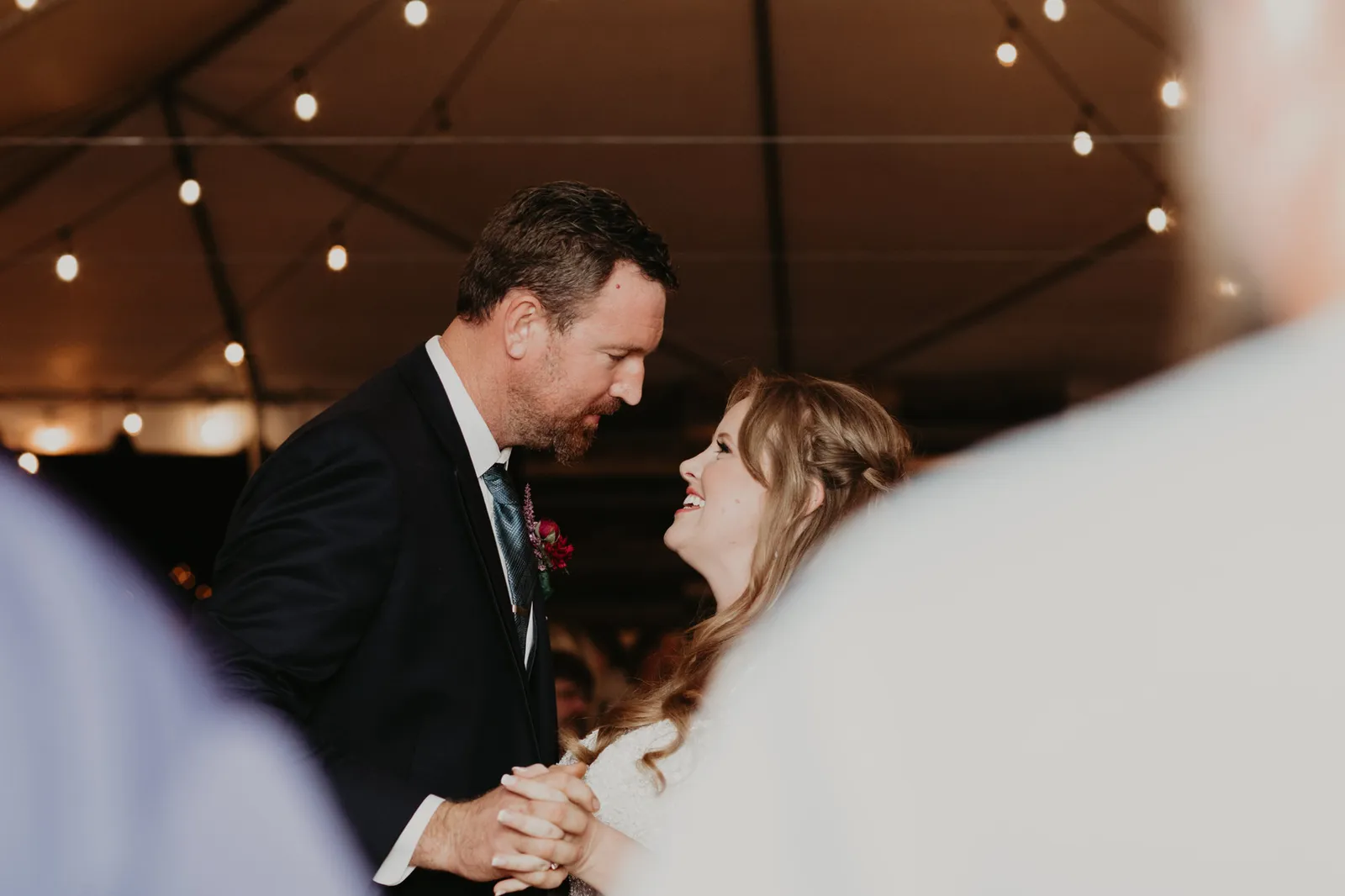 Bride and groom dancing under string lights, seen between guests