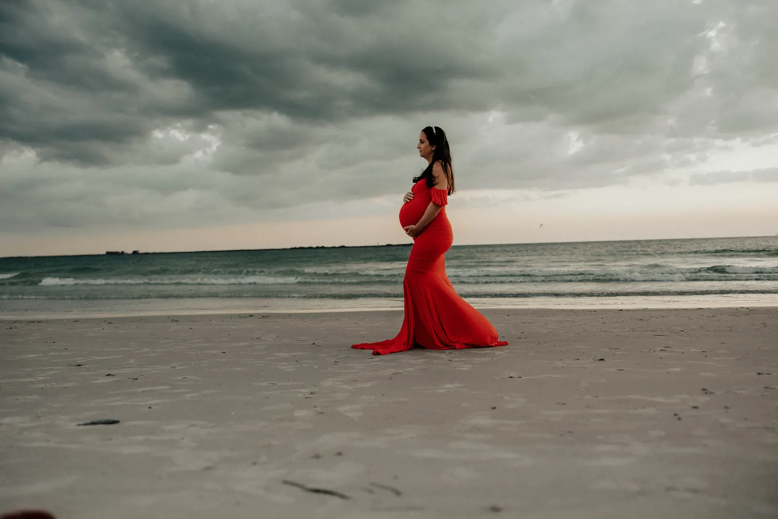 Expectant mother in profile on the open beach, her red train spread on the sand