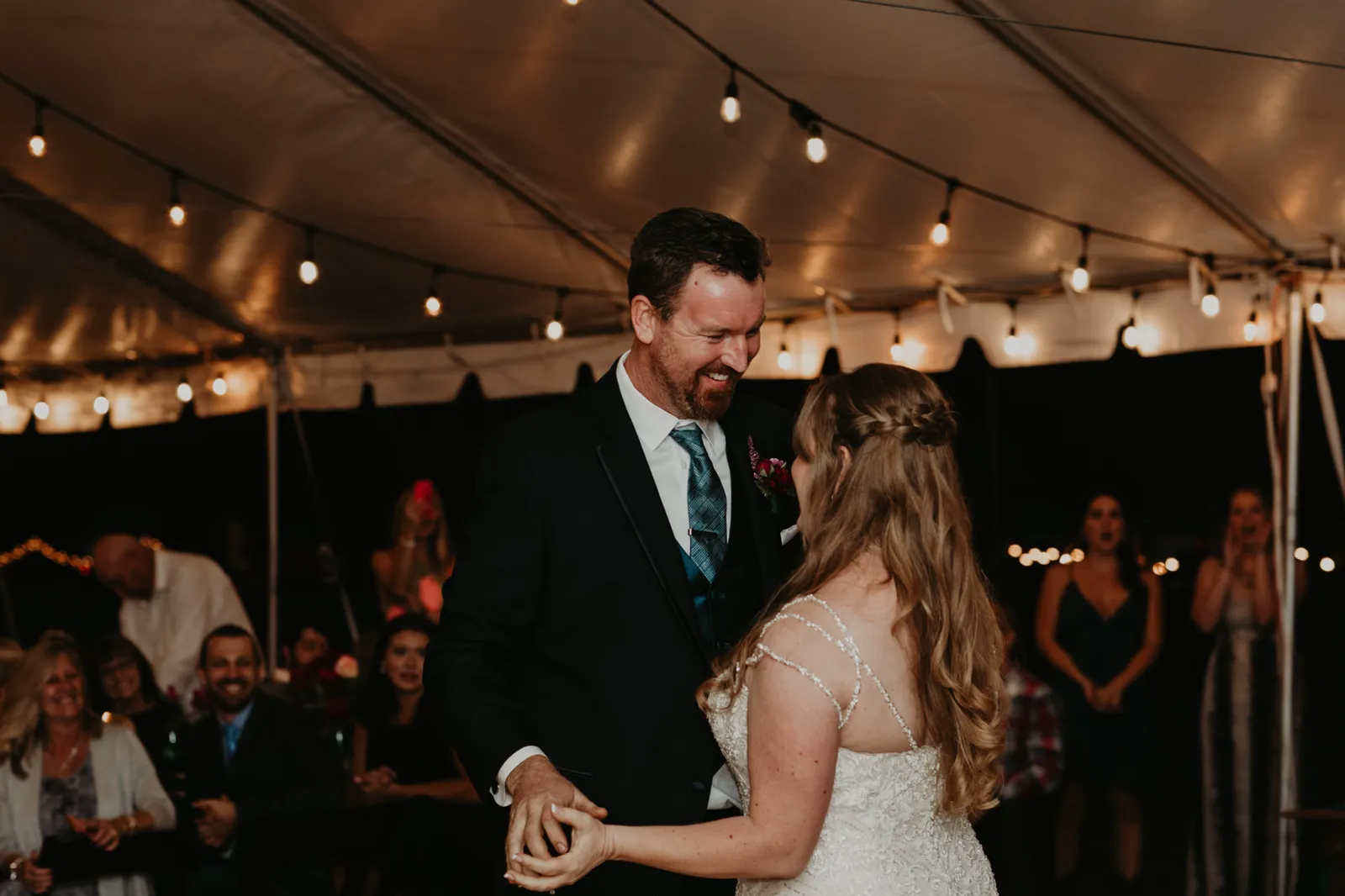 Bride and groom laughing as they dance, guests watching from the edge of the tent