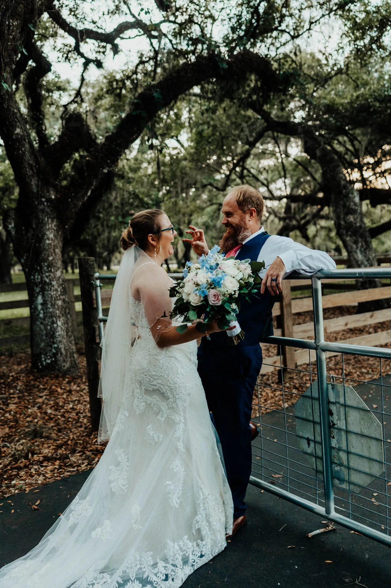 Bride touching the groom's face beside a paddock gate under the oaks