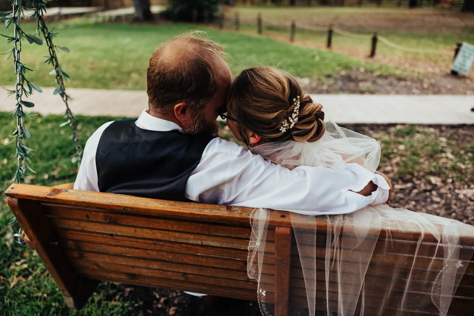 Bride and groom sitting close on a wooden bench swing, seen from behind