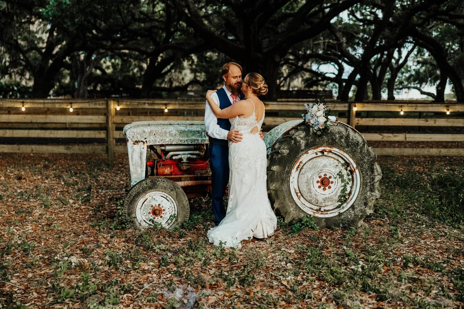 Bride and groom embracing in front of the tractor and a string-lit fence