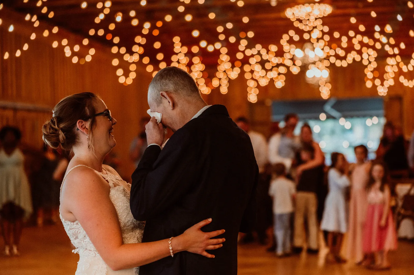 Man wiping his eyes while dancing with the bride under string lights