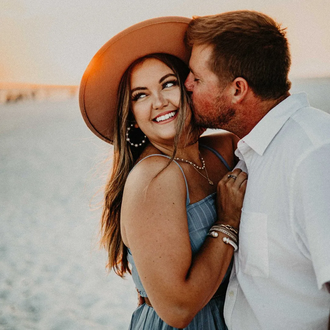 Man kissing a laughing woman in a wide-brimmed hat on the beach
