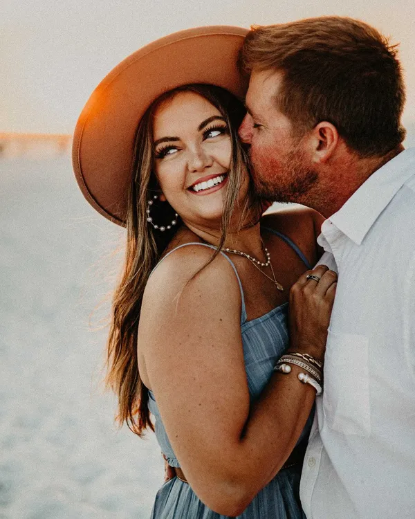 Man kissing a laughing woman in a wide-brimmed hat on the beach
