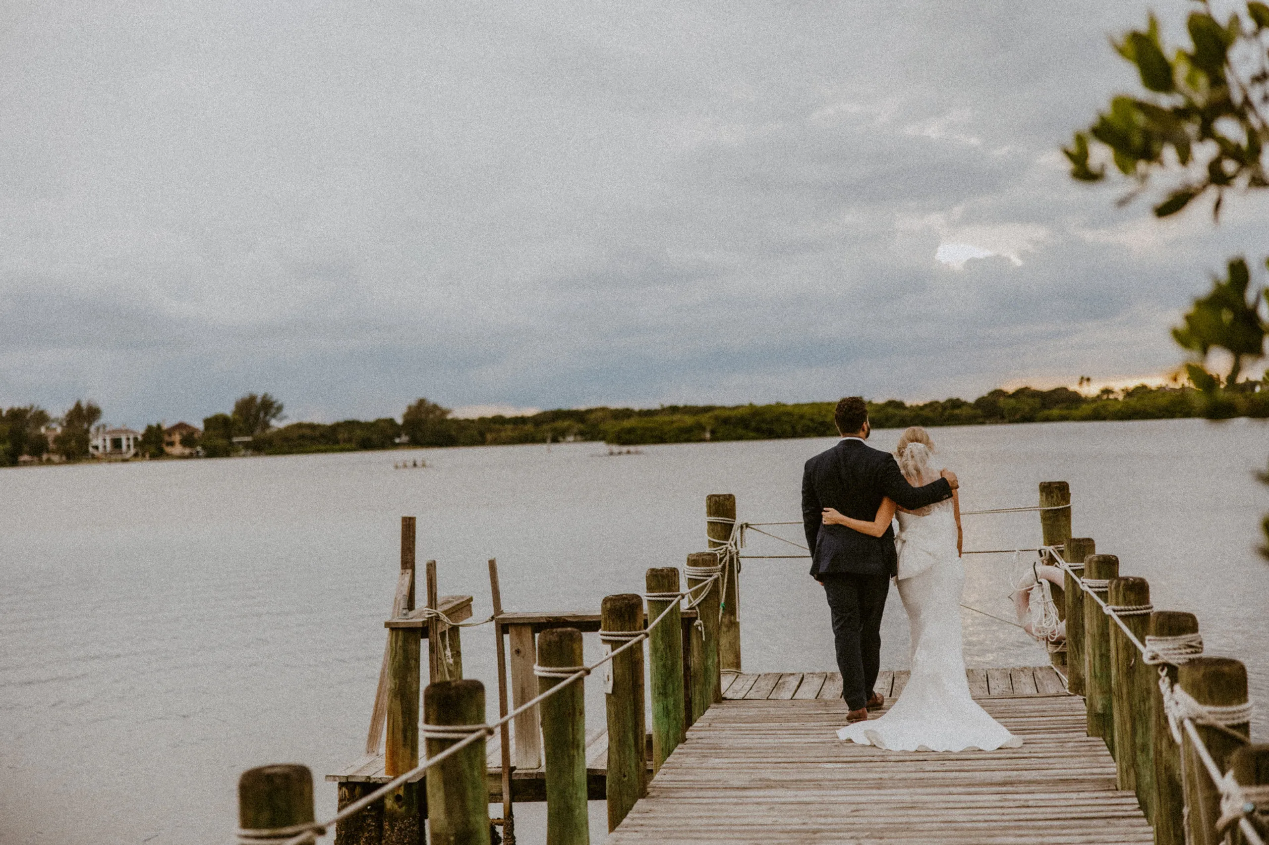 Bride and groom standing arm in arm at the end of a wooden dock, looking out over the water